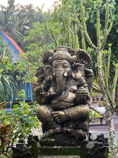A stone statue of Ganesha, the Hindu deity, rests in an outdoor setting surrounded by greenery. The sculpture displays intricate details, with multiple faces and an elephant trunk. The background features lush foliage, including leafy trees, a succulent-like plant with thorny ridges, and a glimpse of a building with a red roof and blue trim. The statue is aged with some moss growth.