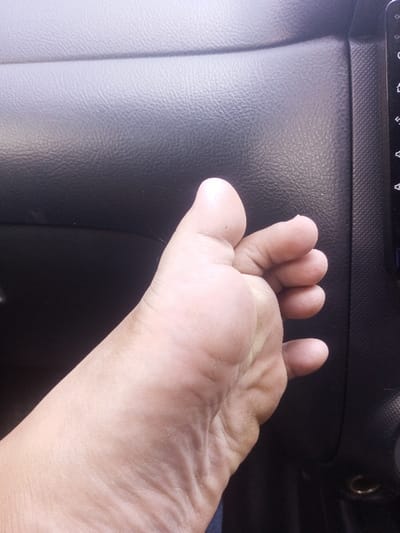 A bare human left foot, seen from its medial side and sole, is prominently positioned within the interior of a vehicle, located in Chori, Nigeria. The skin of the foot is light brown, exhibiting natural creases and minor surface particulate accumulation, consistent with dirt or dry skin, particularly on the heel and sole. The toes are partially flexed. The immediate background consists of a dark gray, textured synthetic material, typical of an automotive dashboard or interior panel. To the right, a darker, vertically oriented panel with a finer texture is visible, bordering the edge of what appears to be an electronic display or control unit with subtle, illuminated symbols. The scene is illuminated by ambient light from an unseen source.