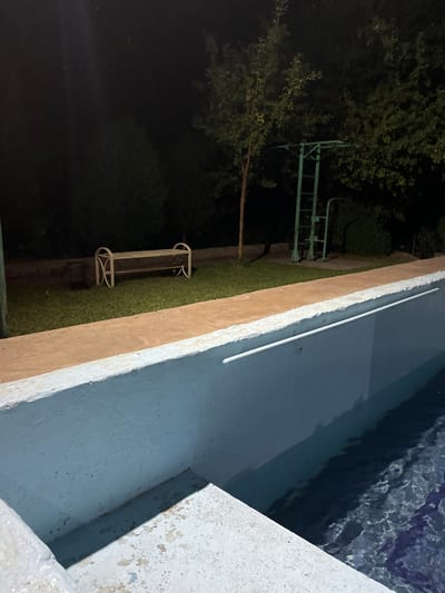 The image depicts an outdoor scene at night, featuring a swimming pool and its surroundings. The pool itself is partially visible, with its light blue interior showing some discoloration and wear. A white handrail is attached to the interior wall of the pool. The pool deck appears to be made of concrete with an orange-colored border.
Beyond the pool, a grassy area is visible, with a wooden bench with a curved back and metal legs positioned on it. To the left of the bench is a dark, cylindrical object, possibly a trash can or a decorative element. In the background, there are trees and a green metal structure that resembles playground equipment or a workout bar. The sky is dark, indicating nighttime, and there are faint specks visible, which could be dust or insects.
The overall impression is that of a slightly neglected outdoor recreational area, possibly a private yard or a small public park, at night. The location is identified as Mashat, Kazakhstan. There are no people visible in the image, and no specific actions or interactions are depicted.