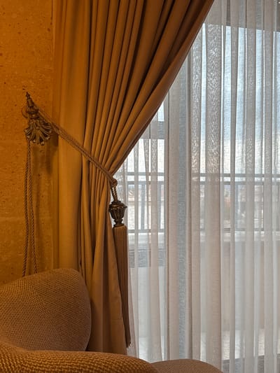 A close-up, indoor shot shows a window with horizontal blinds partially covered by a draped, light-brown curtain. The curtain is held back with a decorative rope and tassel. A chair, upholstered in a similar light-brown fabric with a textured pattern, is partially visible on the right side. The wall, visible at the bottom left, has a rough, textured surface in a beige color. The scene appears to be in a residential setting. The location, Ürgüp, Turkey, is not discernible within the image itself, though it is included within the instructions.