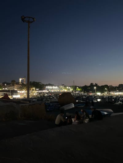 The image depicts a twilight scene in the city of Tipaza, Algeria, likely near the port or a waterfront area. A tall lamppost with multiple lights stands prominently in the left foreground, its wooden pole weathered. In the middle ground, a cluster of people are seated on the ground, appearing to be engaged in conversation or observing the surroundings. One individual on the far right is partially obscured by shadow. Beyond them, a harbor is visible, filled with numerous small boats, some of which are pulled ashore. Buildings with illuminated windows line the waterfront in the background, suggesting commercial activity or residences. A distinct white building with two towers, one featuring a colorful flag, is situated further inland, possibly a landmark. The sky is a deep blue, transitioning to lighter hues near the horizon, indicative of the period just after sunset. The overall atmosphere is calm and subdued, characteristic of an evening in a coastal town.