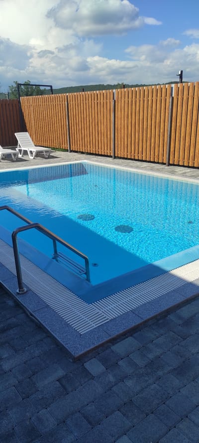 This image displays an outdoor swimming pool with crystal-clear blue water. The pool is surrounded by a paved area made of dark grey interlocking stones, with a wooden slatted fence along one side. The fence is made of horizontal wooden planks and appears to be a light brown color. In the distance, beyond the fence, a lush green landscape with trees is visible, against a bright blue sky filled with white, fluffy clouds.
The water in the pool is inviting and reflects the sky. Several circular features, possibly jets or lights, are visible beneath the surface of the water. A metal handrail leads into the pool, suggesting a step or an entry point. Two white lounge chairs are positioned on the paved area to the left of the pool, indicating a place for relaxation and sunbathing.
The scene suggests a sunny day, likely in the afternoon, given the bright sky and clear visibility. The overall setting appears to be a resort, a private residence, or a recreational area in Tulskiy, Russia. The atmosphere is serene and peaceful, evoking a sense of leisure and enjoyment. There is no visible text in the image.
