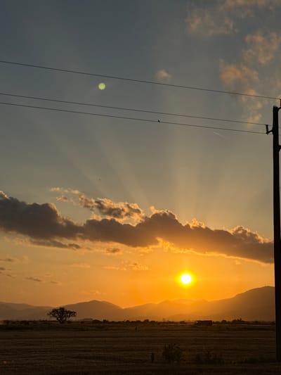 The image captures a sunset over a rural landscape in the vicinity of Blatets, Bulgaria. The sky is a gradient of orange and blue, with rays of sunlight emanating from the setting sun. Scattered clouds are illuminated by the sunset's glow. In the foreground, a dark, cultivated field stretches towards a row of silhouetted mountains. A solitary tree stands in the middle ground. Utility poles with wires are visible in the upper portion of the image. The overall scene conveys a sense of tranquility at dusk.