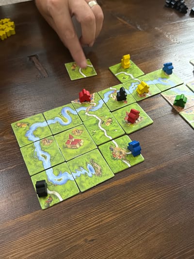 In this overhead shot, a game of Carcassonne is in progress on a dark wooden table. The game board is made up of landscape tiles depicting rivers, fields, and towns. Meeple, which are small wooden player pieces, are placed on some of the tiles, indicating ownership or control. The meeples are in various colors: black, yellow, blue, green, and red. A human hand with a silver ring on the ring finger is visible, actively engaged in the game by placing a tile. The background is out of focus, but the texture of the wooden table is clear. The game is set in the city of İzmir, Turkey.