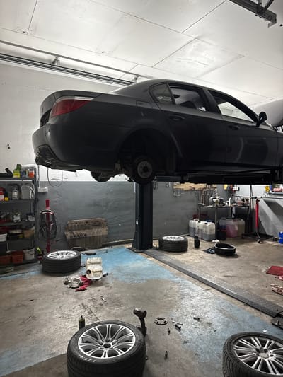 In an auto repair shop located in Čelákovice, Czechia, a dark sedan is elevated on a two-post car lift. The vehicle appears to be undergoing maintenance, as one of its rear wheels is absent, exposing the brake assembly. Several tires are scattered on the concrete floor, with two prominently positioned in the foreground. Various tools and automotive fluids are visible on shelves and the floor, indicating an active workspace. The environment is dimly lit, with overhead fluorescent lights illuminating the scene. The walls are painted a muted gray, and the floor is a combination of concrete and blue epoxy. A large, metallic underbody shield lies on the floor near the car lift, suggesting it may have been removed for inspection or repair.