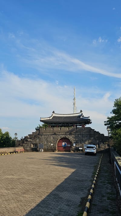 This is a vertical, eye-level shot of the Hwaseong Fortress in Suwon-si, South Korea, under a bright blue sky with scattered white clouds. The fortress is constructed from stone blocks and features a traditional Korean tiled roof structure above a large, arched red gate. A white SUV is parked in front of the gate. To the left of the gate, there is a bench. A tall radio tower is visible in the background, behind the fortress structure. The foreground is paved with interlocking bricks, and a line of yellow and black striped bollards runs along the right side of the paved area. Lush green trees are visible on the left and right sides of the fortress.