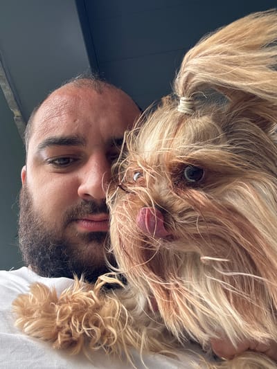 A close-up selfie taken outdoors shows a man holding a small, light-brown dog. The man, positioned on the left, has a beard and is looking down at the dog, his left eye visible. The dog, occupying the right side of the frame, has its tongue out, licking its nose. The dog's fur is long and somewhat messy, with a ponytail secured with a light-colored band. The background consists of a dark, textured surface, possibly a ceiling or overhang. The image is taken from a low angle, suggesting the man is looking upwards. No specific landmarks of Epping Forest, United Kingdom, are identifiable.
