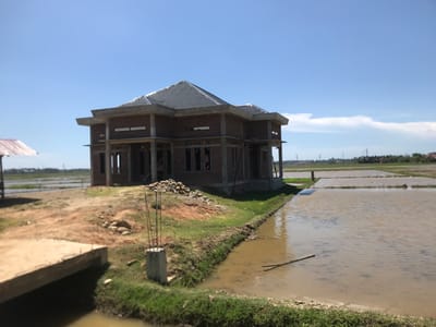 The image depicts a two-story brick house under construction in Uleegampong, Indonesia. The house has a grey tile roof and concrete framing. The building site is adjacent to flooded rice paddies. A pile of stones sits near the house. In the foreground, there is a concrete slab and a concrete pillar with rebar extending upwards. The sky is clear with some clouds on the horizon. Utility poles and distant buildings can be seen in the background.