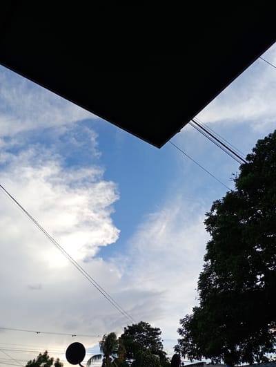 View looking upwards in Barquisimeto, Venezuela. A dark rectangular overhang covers the top of the frame. Below, a cloudy blue sky is partially visible. Power lines cut diagonally across the scene. In the lower right, the canopy of a large tree occupies the space. Below, smaller trees and a utility pole are partially visible. The location is discernable due to the distinctive architectural style and vegetation common to the region.
