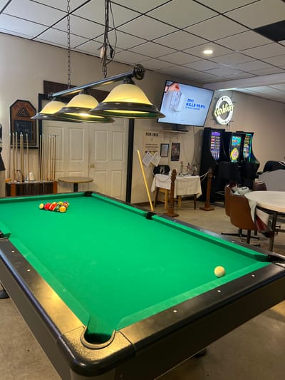The image depicts the interior of a bar or recreational establishment, specifically a pool hall, located in Atlantic Beach, United States of America. A green pool table with a rack of colored balls is prominently featured in the foreground. One white cue ball is visible on the table. Above the pool table, a three-bulb chandelier with yellow shades hangs from the ceiling. In the background, there are several slot machines, a television screen displaying an advertisement for "zevo KILLS 99.9%," and a neon sign that reads "carblin." A round table with chairs is also visible, suggesting seating areas. The overall environment appears casual and recreational, with a tiled ceiling and plain walls. There are no people clearly visible in the image.
