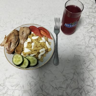 A plate of food sits on a white, textured surface, next to a glass of red liquid and a fork. The plate contains several pieces of cooked chicken, a portion of what appears to be noodles or pasta with a creamy sauce drizzled over it, slices of tomato, and several slices of cucumber. The chicken pieces are golden brown and appear to be roasted or baked. The pasta is a light yellow color, and the sauce is white and thick. The tomatoes are bright red, and the cucumber slices are green with visible seeds.
The glass next to the plate is filled with a dark red beverage, and a Nestlé logo is visible on the side of the glass. The fork is silver and has four tines. The white surface beneath the plate and glass has a marbled or crumpled texture. The lighting suggests it is daytime, likely indoors. The image is a top-down view of the meal, with the food taking up the majority of the frame. The overall impression is of a home-cooked meal.