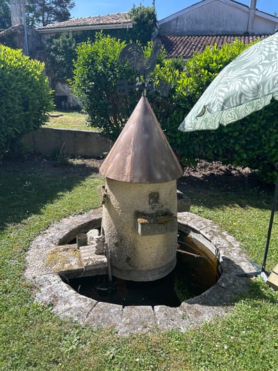 A stone well with a conical metal roof sits in a grassy yard in Saint-Germain-de-Lusignan, France. The well is cylindrical, made of aged stone, with some visible moss and possible algae growth. A stone surround forms the well's opening, with what looks like a pipe sticking out from the side of the well. The metal roof has a weather vane affixed to it. An umbrella with a light green and white leaf pattern is partially visible in the background, along with bushes and a building. The yard is green with short grass.