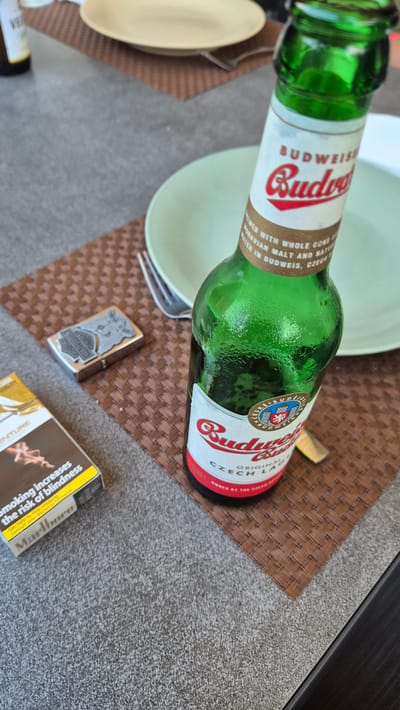A close-up, high-angle shot depicts a table setting outdoors, possibly in Dissen am Teutoburger Wald, Germany. A green glass bottle of Budweiser beer, showing condensation, stands prominently on a brown woven placemat. The label reads "BUDWEISER Budweiser CZECH LAGER". Next to the beer bottle, on the left, is a pack of Marlboro cigarettes and a silver Zippo lighter. A fork rests on the placemat near the beer bottle. In the background, two plates are visible: a light green one and a beige one. The overall setting suggests a casual outdoor meal or break.