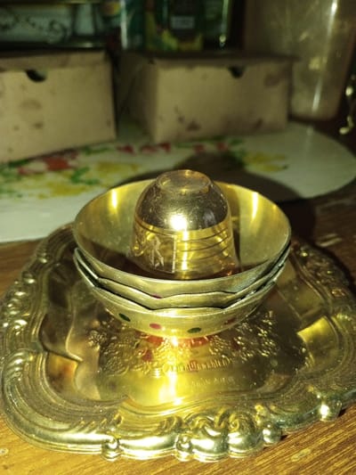 A close-up photograph taken indoors in Longmiang, Laos, displays a still life arrangement of brass-colored items on a wooden surface. A rectangular, ornate brass-colored tray with raised scalloped edges and embossed patterns forms the base. The inscription "MADE IN INDIA" is partially visible on its surface. Stacked centrally on the tray are three brass-colored bowls. The lowest visible bowl features small red and green circular accents on its exterior rim. Inverted within the uppermost bowl is a small, conical, brass-colored object, possibly a thimble or miniature cup. It exhibits multiple horizontal etched bands and a distinct, stylized emblem near its broader base. The background, out of focus, includes a patterned textile surface displaying red, yellow, and green elements, situated behind two light brown cardboard boxes with cut-out handles. Other indistinct items, potentially bottles, are also present. The lighting is warm and indirect, revealing minor dust accumulation on the metallic surfaces.