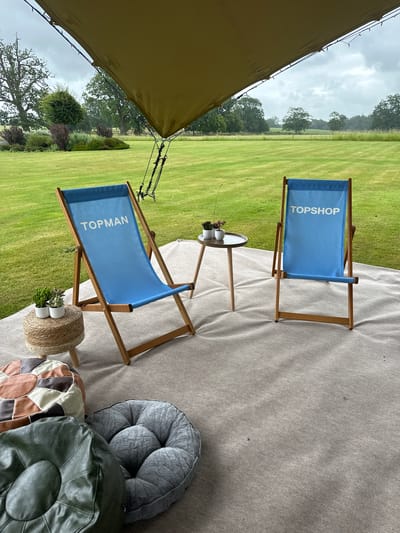 A scene of two blue deck chairs with white text that reads "TOPMAN" and "TOPSHOP" is captured outdoors. The chairs are set up on a neutral-colored rug, with a small round table and two small potted plants placed between them. In the foreground, there are several floor cushions in various colors and textures, including brown and cream patchwork, dark green leather, and grey. Behind the chairs, a lush green lawn stretches out towards a treeline under a cloudy sky. The setup appears to be under a large, tan-colored awning supported by poles and ropes. The overall impression is a comfortable outdoor lounge area.