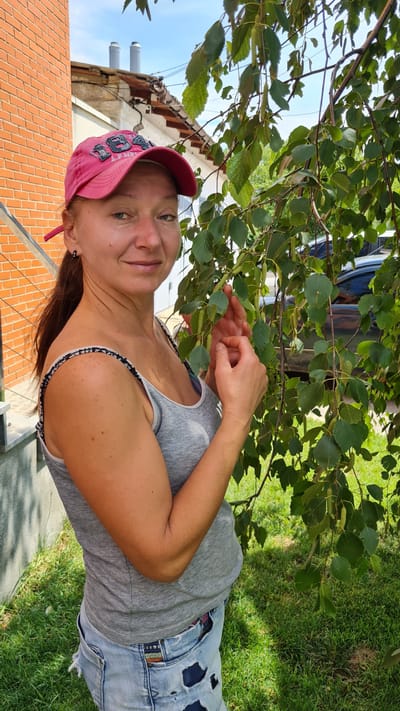 A woman wearing a pink baseball cap and a grey tank top stands outdoors, partially obscured by tree branches. She is also wearing ripped denim shorts. Her right arm is bent, and her hand is near the leaves of the tree. She smiles at the camera. The scene appears to be a sunny day in a residential area, with a brick building visible in the background. The location is Shabo, Ukraine. There is visible text on the woman's cap, but it is difficult to read clearly.