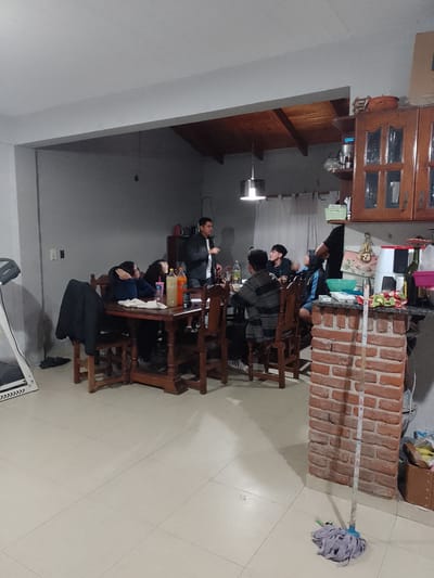 The image depicts a gathering inside a residence, likely in Gobernador Julio A. Costa, Argentina. Several people are seated around a wooden dining table. Some are leaning on the table, while one man is standing and appears to be speaking or gesturing. Food and beverages are present on the table. To the right of the table, a brick-faced counter or support column is visible with kitchen items on top. A mop rests on the tiled floor next to the brick structure. In the background, there is wooden cabinetry and a hanging light fixture. A treadmill is visible in the left background. The overall lighting suggests an indoor setting, possibly in the evening.