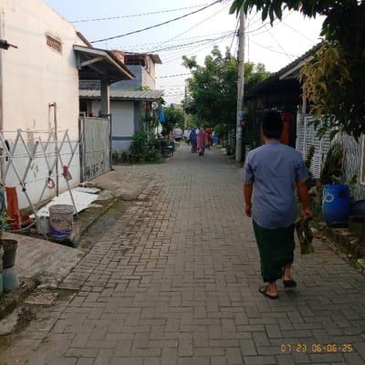The image shows a brick-paved alleyway in the city of Kecamatan Panongan, Indonesia, located at the coordinates -6.276 latitude and 106.551 longitude. There are several houses along the alleyway, with a white wall on the left side of the image. A man in a blue shirt and a green sarong is walking towards the camera. He is holding a green cloth in his right hand. Three women are walking away from the camera in the distance. The image is taken on a sunny day, with a blue sky in the background. There is a blue bucket visible in the lower right of the image. A white laundry rack is located on the left side of the image.