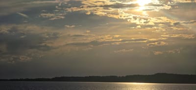 A wide shot captures a serene natural landscape with a vast body of water in the foreground. The water reflects the bright, golden sunlight that pierces through scattered, textured clouds. The sky above is a dramatic canvas of light and shadow, with the sun positioned in the upper right quadrant, casting radiant beams. A dark, silhouetted treeline forms the horizon, suggesting land beyond the water, with indistinct structures or power lines visible on the distant hills. The overall scene evokes a sense of tranquility and the beauty of a sunset or sunrise over a peaceful, natural expanse. The specific location is stated to be the city of Pristan'-Przheval'sk, Kyrgyzstan.