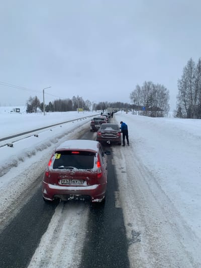 The image depicts a winter scene of a traffic queue on a two-lane asphalt road in Bolgury, Russia. The road surface is partially cleared, revealing dark asphalt interspersed with patches of slush and snow, particularly on the shoulders and between lanes. Snowbanks line both sides of the road, and the surrounding terrain, including sparse trees and fields, is covered in snow under an overcast, light grey sky.

In the foreground, a red Lada Kalina hatchback is visible from the rear, covered in light snow. It bears Russian license plate Y 635 KY 190 and has a yellow exclamation mark sticker on its rear window. Its taillights appear illuminated. Immediately ahead of this vehicle is a dark-colored sedan, possibly brown or bronze, with Russian license plate O 750 PO 190.

A male individual, dressed in a dark blue jacket and dark trousers, is bending down at the rear driver's side of the dark-colored sedan, appearing to interact with the vehicle or the ground near its rear tire. Further down the road, a line of approximately six to eight additional vehicles, including sedans and potentially larger trucks or vans, are stopped or moving slowly. Some of these vehicles are also snow-dusted. Utility poles with power lines and a road sign indicating a blue square with a white arrow are visible on the right side of the road in the midground. The entire scene indicates cold, snowy conditions and a vehicular stoppage or slow movement.