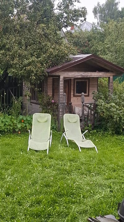 In the garden of a dacha in Orbita SNT (Terpigoryevo), Russia, two empty, light green deck chairs are positioned on a grassy lawn in front of a small wooden shed. The shed has a brown roof and light-colored wooden siding, with an open doorway and a window visible. Lush green foliage, including trees and bushes, surrounds the shed and the yard. The ground is covered in thick, uneven grass, with some smaller white wildflowers scattered throughout. The sky is overcast, contributing to the subdued lighting of the scene.