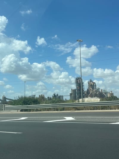 The image shows a view from a car on a highway. The foreground is the asphalt road with white lane markings. A metal guardrail separates the highway from the adjacent area. Beyond the guardrail, there is vegetation and an industrial complex with various structures, tanks, and pipes. In the background, a blue sky with scattered white clouds is visible, with power lines running across the sky. Based on the presence of the industrial complex and the highway, the image may be located in Ramla, Israel.