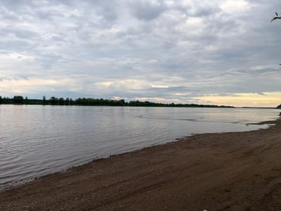 This is a horizontal, eye-level photograph of a wide, calm river with a sandy and pebbly shore in the foreground. The sky is overcast with numerous gray clouds, some of which are tinged with yellow and orange at the horizon, suggesting it may be sunrise or sunset. The water is a muted gray-blue and reflects the sky. A dense line of green trees borders the opposite bank of the river, stretching across the horizon. The shore is composed of dark, damp earth mixed with various-sized pebbles and some tire tracks. There are no people or objects clearly visible on the shore or in the water, though a dark silhouette that might be a person is visible on the far right bank. The scene evokes a sense of tranquility and natural beauty under a cloudy sky. The location is identified as Yaromaska, Russia. No text is visible in the image.