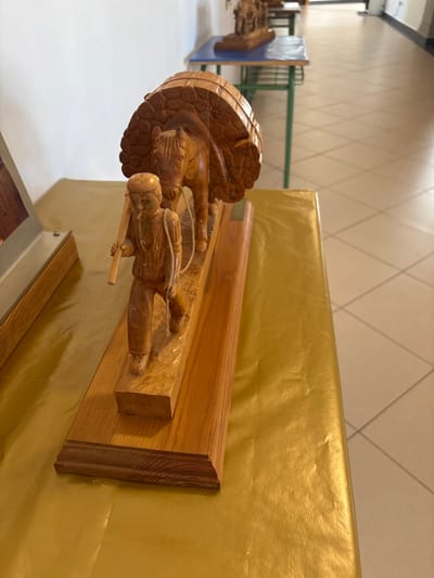 The image displays a finely crafted wooden sculpture situated in an indoor exhibition space in El Espinar, Spain. The primary subject is a light-brown wooden carving on a rectangular wooden base, depicting a male figure walking ahead of a horse. The man wears a cap and trousers, carrying a long staff or tool over his right shoulder. The horse, following the man, bears a large, rounded, textured load on its back, resembling a stack of agricultural goods such as hay bales, secured by visible lines.
The sculpture rests on a surface covered with a gold-colored, slightly wrinkled material. To the immediate left, a portion of another wooden display item with a metal top edge is visible. The background reveals a clean, well-lit hallway with a plain white wall on the left and a light-colored, square-tiled floor extending into the distance on the right. Further down the hallway, additional similar wooden sculptures are visible, displayed on small tables with light-blue tops and green metal frames. Natural or artificial light illuminates the scene, casting subtle shadows.