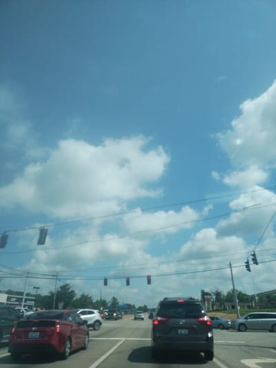 This media file, captured in Nicholasville, United States of America, presents a view of a roadway with a clear blue sky and scattered clouds. The image appears to be taken from inside a vehicle, looking outwards.
The primary subjects are several cars lined up on a road, suggesting a traffic scenario or perhaps a parking lot. In the foreground, a dark-colored SUV is visible, with its brake lights illuminated. Further down the road, a red car and a white car are also present. Overhead, traffic lights are mounted on poles, with some displaying red lights. Telephone wires crisscross the sky, interspersed with fluffy white clouds.
The scene suggests it is daytime, likely late morning or afternoon, given the bright sky. The weather appears to be fair. While no people are clearly discernible as subjects, the presence of vehicles indicates human activity. The setting is clearly an outdoor, urban or suburban environment with infrastructure typical of a road or commercial area. No discernible text is present in the image.