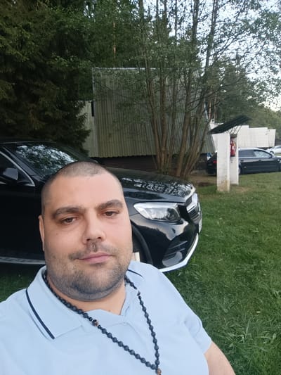 The image depicts a man posing for a selfie outdoors. He is positioned in the foreground, with a black car partially visible behind him. In the background, there are trees and some structures that appear to be buildings with corrugated metal siding. One of the structures has a visible fire extinguisher mounted on a white pillar. The ground is covered in green grass. The man is wearing a light blue polo shirt and a dark beaded necklace. The location is identified as the city of Babylon, Czechia.