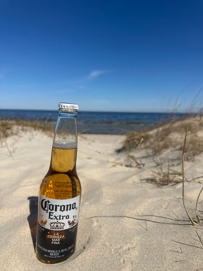 A bottle of Corona Extra beer lies on a sandy beach. The beer bottle is oriented horizontally, with the bottle cap facing towards the sky. The background consists of the sea and sky. The sand appears to be coarse and light-colored. There are some dry, thin blades of grass near the beer bottle. The image does not depict Zīvartiņš, Latvia.