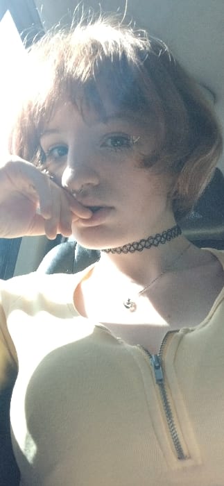 A frontal portrait depicts an individual with short, reddish-brown hair and bangs. The individual has light skin, blue eyes, and a small silver hoop piercing in the right nostril. Their right hand, specifically the thumb and index finger, is positioned near the lower lip. The individual wears a light yellow garment with a silver zipper extending down the front. A black choker necklace with an interlocking pattern is around their neck, and a thin silver chain with a small, dark-colored pendant hangs below it. The background is indistinct and dark, consistent with an indoor or vehicle interior setting. Strong backlighting from the upper left illuminates the individual's hair and creates highlights on their face and clothing. The image contains no discernible features to indicate the specific location in the city of Bistrice, Montenegro.