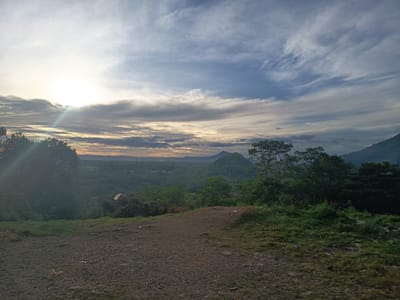 This is an outdoor landscape image captured in Geundring, Indonesia, depicting a panoramic view likely during sunrise or sunset, evidenced by the low-angle sun emanating a prominent lens flare from the upper-left quadrant. The upper portion of the frame features a sky with layered clouds, ranging from wispy cirrus-like formations in the lighter blue areas to denser, greyish stratiform clouds across the mid-sky. The sun's intense light creates a bright, almost blown-out area in the upper left, casting a soft, warm glow onto the distant horizon where the sky transitions into muted orange and yellow hues.
The midground showcases a vast expanse of lush, dense green vegetation, predominantly trees, suggesting a forested valley or plain. In the central-right midground, a prominent, conical mountain, heavily covered in dark green foliage, rises distinctly against the hazy distant landscape. Further to the right, faint outlines of additional, less defined hills or mountains are visible. A small, reddish-brown structure, possibly a rooftop, is partially discernible amidst the trees in the midground, slightly to the left of the central mountain.
The foreground consists of a relatively flat, textured dirt path or clearing, marked by variations in brownish-grey earth and scattered small rocks. Patches of sparse, light green and dry yellowish grass are interspersed across this area, particularly on the right side. Thick, dark green foliage, including large trees, frames the left side of the foreground, while abundant green bushes and grass occupy the right. Small pieces of visible debris, including white and blue plastic, are scattered on the ground in the immediate foreground. No people or animals are present in the scene.