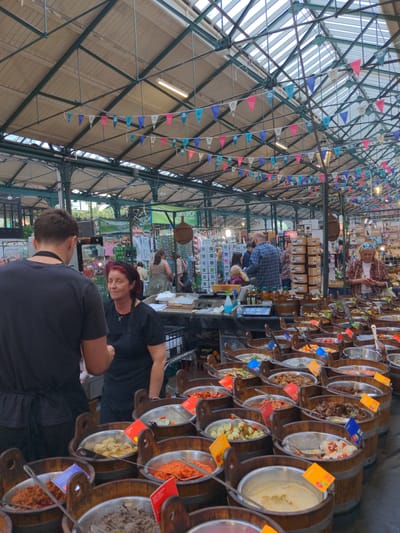 The image captures a bustling indoor market scene, likely located at St George's Market in Belfast, United Kingdom, given the visible signage. The market features numerous food stalls, with a prominent display of wooden barrels filled with various food items, possibly olives, relishes, or dips, each with descriptive labels. In the foreground, a person wearing a black apron with red hair is seen interacting with another person obscured from view, likely a customer or staff member. Behind them, other shoppers and vendors are visible, browsing stalls laden with produce and other goods. The market has a covered ceiling with exposed metal beams and a translucent roof, allowing natural light to filter through, creating a vibrant and open atmosphere, further enhanced by strings of colorful triangular flags draped across the ceiling.