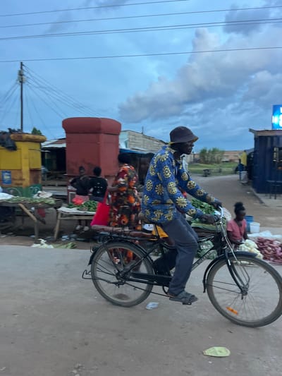 In Kapiri Mposhi, Zambia, a man wearing a blue, patterned shirt and a cap rides a black bicycle loaded with goods on a paved road adjacent to a market area. The market includes various stalls selling produce such as onions and green vegetables. Other people are present in the market area. Overhead power lines are visible against a cloudy sky.