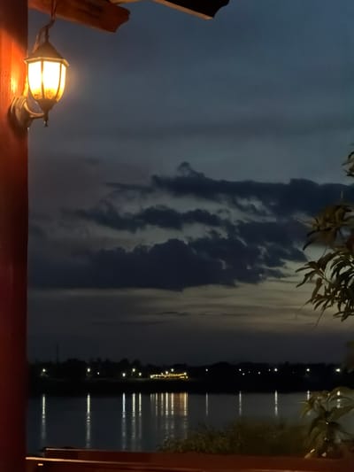 An outdoor scene at dusk shows a lit lamp on the left side, casting a warm glow on a dark red pillar and the wooden structure above. The lamp's glass enclosure is visible. Beyond the lamp, the sky is a deep blue-gray with dark, textured clouds. In the middle ground, a dark silhouette of a treeline and buildings is visible across a body of water, punctuated by numerous small, bright lights. The water reflects these lights as vertical streaks. The foreground shows a wooden railing. The foliage of a plant is visible on the far right, silhouetted against the sky. The scene appears to be from a vantage point overlooking the Mekong River in Vientiane Capital, Laos, possibly a restaurant or cafe with a river view.