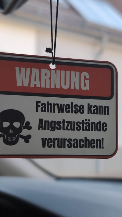 The image shows a close-up of a warning sign hanging from a string. The sign is red and white with black text and a skull and crossbones graphic. The text on the sign reads "WARNUNG" at the top, and below that, "Fahrweise kann Angstzustände verursachen!". This translates to "WARNING: Driving style can cause anxiety!". The sign appears to be an air freshener or a novelty item. The background is blurred, suggesting the photo was taken from inside a vehicle, possibly in Bingen am Rhein, Germany, given the German text on the sign. The lighting suggests it is daytime, and the background colors indicate an outdoor setting with buildings and possibly sky. There are no people visible in the image.