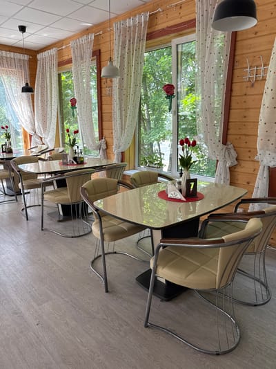 The image shows a brightly lit, rustic dining area with wooden paneled walls and large windows looking out onto a green, leafy exterior. Several tables are set with light-colored chairs. The tables have reflective, possibly glass or laminate, surfaces. Each table has a vase of red tulips, napkins, and what appear to be salt and pepper shakers or condiment bottles. A numbered stand, possibly indicating table number 7, is visible on one table. The chairs have a cream-colored padded seat and back, with a chrome wire frame. The flooring is light-colored wood laminate. Two pendant lights hang from the white tiled ceiling. The overall impression is of a clean and tidy restaurant or cafe interior. The location is identified as Серово, Russia.