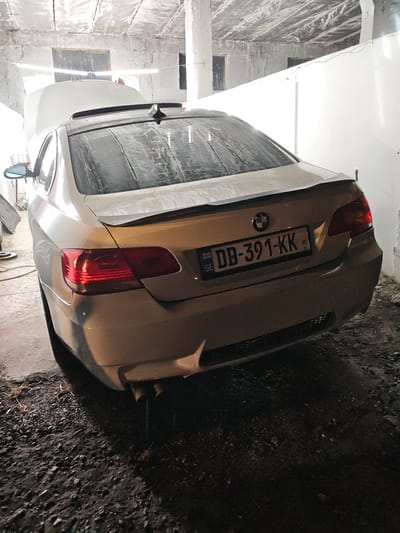 A rear three-quarter view shows a light-colored BMW coupe parked indoors on a gravel floor, possibly in a garage or workshop, in Krtsanisi, Georgia. The car's trunk is open, revealing its interior. The license plate is visible and reads "DB-391-KK GE". The interior of the structure features rough concrete walls and ceiling. The lighting is dim, with a bright light source coming from above and to the left. The ground is covered in loose gravel and some debris. There are no people visible in the image.