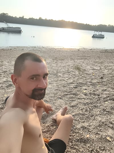This is a selfie taken at Dunakeszi, Hungary, on a beach next to a river. The subject is a man with short brown hair and a beard, wearing black shorts. He is sitting on the pebble beach, looking at the camera with a relaxed expression. In the background, the river is calm, reflecting the light of the setting sun. There are two pontoon boats on the water, one further in the distance and another closer to the shore. The trees lining the opposite bank are silhouetted against the bright sky. The time of day is late afternoon or early evening, as indicated by the warm, golden light and long shadows. The weather appears to be pleasant and clear. The overall mood of the image is peaceful and serene. There is no visible text in the image.