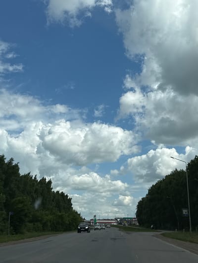 The image depicts a highway scene in Nurkeevo, Russia, under a partly cloudy sky. The road, paved with asphalt, curves slightly to the right. A line of dark green trees flanks both sides of the road, with the trees on the left appearing denser and taller. Several cars are visible on the highway, traveling in the same direction as the viewer. On the left side of the road, a blue street sign with the number "77" is present. In the distance, a commercial area with gas stations and buildings can be observed. A street light stands on the right side of the road, near the edge of the trees. The sky is a mix of bright blue and large, white and gray clouds.