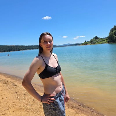 A woman stands on a sandy beach with a calm, blue lake behind her. She is wearing a black bikini top and patterned swim trunks. Her left hand is on her hip, and her right arm is bent. The background shows a tree-lined shore and hills in the distance under a clear blue sky with a few scattered clouds. There are some people on paddleboards in the lake. The location is the city of Cherry Grove, United States.