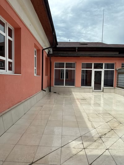 The image presents an outdoor architectural scene, likely a courtyard or side of a building, under an overcast daytime sky. The primary subjects are a building with a salmon-pink facade and a large tiled surface.

The building features a textured, salmon-pink exterior with numerous white-framed windows of different sizes, some with multiple panes. One window on an upper floor is slightly open. Above the uppermost windows, a section is fitted with glass blocks. The building's roof has dark brown eaves and trim, with black gutters and downspouts running along the facade. A tall, slender lightning rod extends from the roofline upwards into the sky.

Adjacent to the building, a large flat area is covered with light-colored, square tiles, appearing beige or off-white. These tiles exhibit signs of wear, dirt, and streaking, particularly noticeable along the dark grout lines. Some sections of the tiled surface show faint diagonal patterns.

The sky is cloudy and overcast, suggesting a diffused light during the day. No human activity or individuals are present in the scene. There is no visible text in the image. The location is Belovo, Bulgaria.