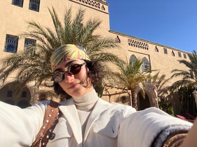 The image is a selfie taken outdoors in Rissani, Morocco, on a clear, sunny day. An adult female subject, approximately 20-40 years old, is positioned in the foreground, facing the camera with a faint smile. She has short, dark, curly hair and wears dark, oval-shaped sunglasses. A vibrant, multi-colored headscarf, featuring patterns in yellow, green, and pink, covers her head. She is dressed in a light-colored, ribbed turtleneck sweater under a light-colored jacket. A brown shoulder bag with silver stud and fringe detailing is visible over her right shoulder. Gold-colored bracelets are worn on her left wrist, which is holding the camera. The background features a large, sand-colored building constructed in a traditional Moroccan architectural style, including intricate arched and rectangular latticework windows, green decorative tile trim along the roofline, and a prominent minaret-like structure on the left side. Several tall palm trees are visible around the building, casting distinct shadows from the bright sunlight. Red outdoor lounge chairs are partially discernible in the far background.