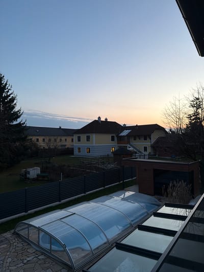 The image presents an elevated view of a residential landscape in Ferschnitz, Austria, during either dawn or dusk, characterized by a gradient sky transitioning from light blue to orange-yellow at the horizon. In the immediate foreground, the edge of a modern structure with dark-framed skylights is partially visible from the upper right, reflecting the sky. Adjacent to this, a stone-paved patio area leads to a swimming pool, which is covered by a clear, arched, retractable enclosure, revealing water beneath.

A dark gray horizontal slatted fence bisects the midground, separating the foreground property from the adjacent ones. Beyond the fence, a maintained lawn area contains various elements: to the left, white industrial containers are stacked near garden plots; to the right, a modern, dark brown, wood-paneled utility building or shed with a flat roof is present, fronted by sparse vegetation. Further back in the garden, a small, dark-roofed gazebo is visible.

In the background, multiple residential buildings are arranged. A large, two-story, light yellow building with a dark gabled roof and numerous dark windows dominates the left-center. To its right, a two-story house features a brown gabled roof, light yellow upper story, and light blue lower story, with internal lights illuminated in at least one ground-floor window. This house appears connected to or adjacent to another structure with a brown roof and yellow walls, potentially via an enclosed glass sunroom or a structure with an external staircase. A large evergreen tree is partially visible on the far left, while bare deciduous trees are visible on the far right. No individuals or active interactions are depicted within the scene.