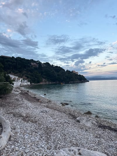 This is a wide-angle, eye-level shot of a pebble beach and coastline in the city of Kassopaia Municipal Unit, Greece, during twilight. The sky is a soft blue with scattered grey and white clouds, tinged with hints of pink and orange from the setting sun. The sea is calm and reflects the muted colors of the sky.

On the left side of the frame, a dense green hillside rises steeply, covered in trees and dotted with houses. White buildings with red-tiled roofs are nestled along the base of the hill, close to the water's edge. A small beach extends along the foreground, composed of grey and white pebbles. A large piece of driftwood lies on the left side of the beach.

In the middle distance, a few small yellow buoys are visible in the water, and two yellow kayaks or paddleboards are occupied by individuals. A thin spit of land juts out into the sea on the right side of the frame, also covered in trees, with a large villa perched on its summit. The overall atmosphere is serene and peaceful, indicative of a quiet coastal evening.