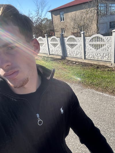 A selfie is taken from a low angle. Part of the person's face, including the ear, forehead, nose, and mouth, is visible on the left. The person wears a dark zip-up jacket. The background features a light gray building with a red roof behind a decorative concrete fence. A strip of grass runs alongside the fence. A paved area is visible on the right side. The sun is out.