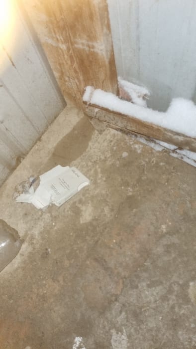 An upward angled view shows a dilapidated space, possibly an attic or basement. Exposed wood beams are visible, some with snow accumulation. The floor is a rough, stained concrete. A torn, partially obscured piece of white paper or cardboard lies on the floor, potentially with printed text. The overall impression is one of disrepair and neglect.