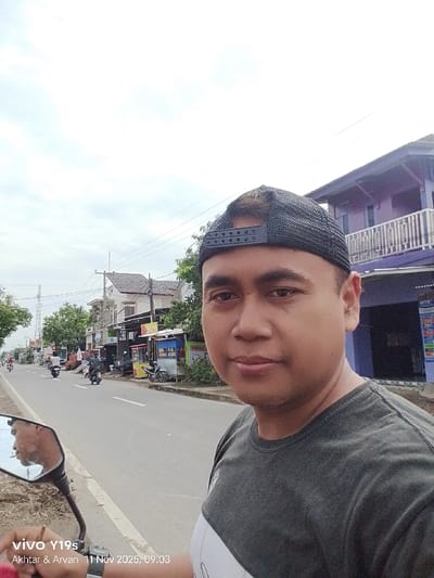 A man wearing a baseball cap is standing on a road, facing the camera. Behind him are buildings, trees, and other motorcycles. There's also a sign on the lower left side of the image, and the reflection of a man in the side mirror. The sky is overcast.