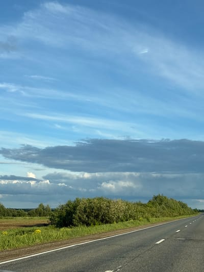 The image shows a rural road scene, likely taken from a moving vehicle. A paved road with white lane markings stretches into the distance on the right side of the frame. Adjacent to the road is a grassy shoulder and then a line of trees and bushes. The sky occupies a significant portion of the image, featuring a mix of light blue sky with wispy clouds and darker, more ominous storm clouds. The location is Kvarsa, Russia. No people are visible. There are no specific actions or interactions to describe. The focus is on the environment: road, vegetation, and sky.